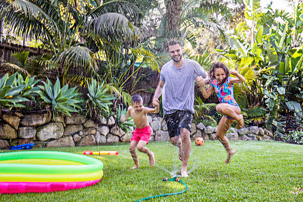 Father and children jumping in sprinkler Father and children jumping in sprinkler in the backyard garden. summer fun stock pictures, royalty-free photos & images